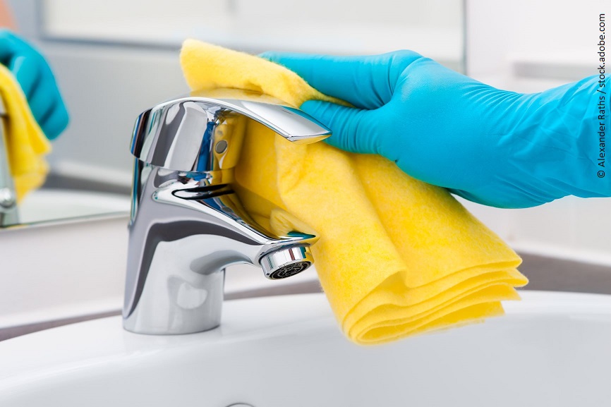 Eine Hand in Putzhandschuhen wischt mit einem Lappen über einen Wasserhahn. 